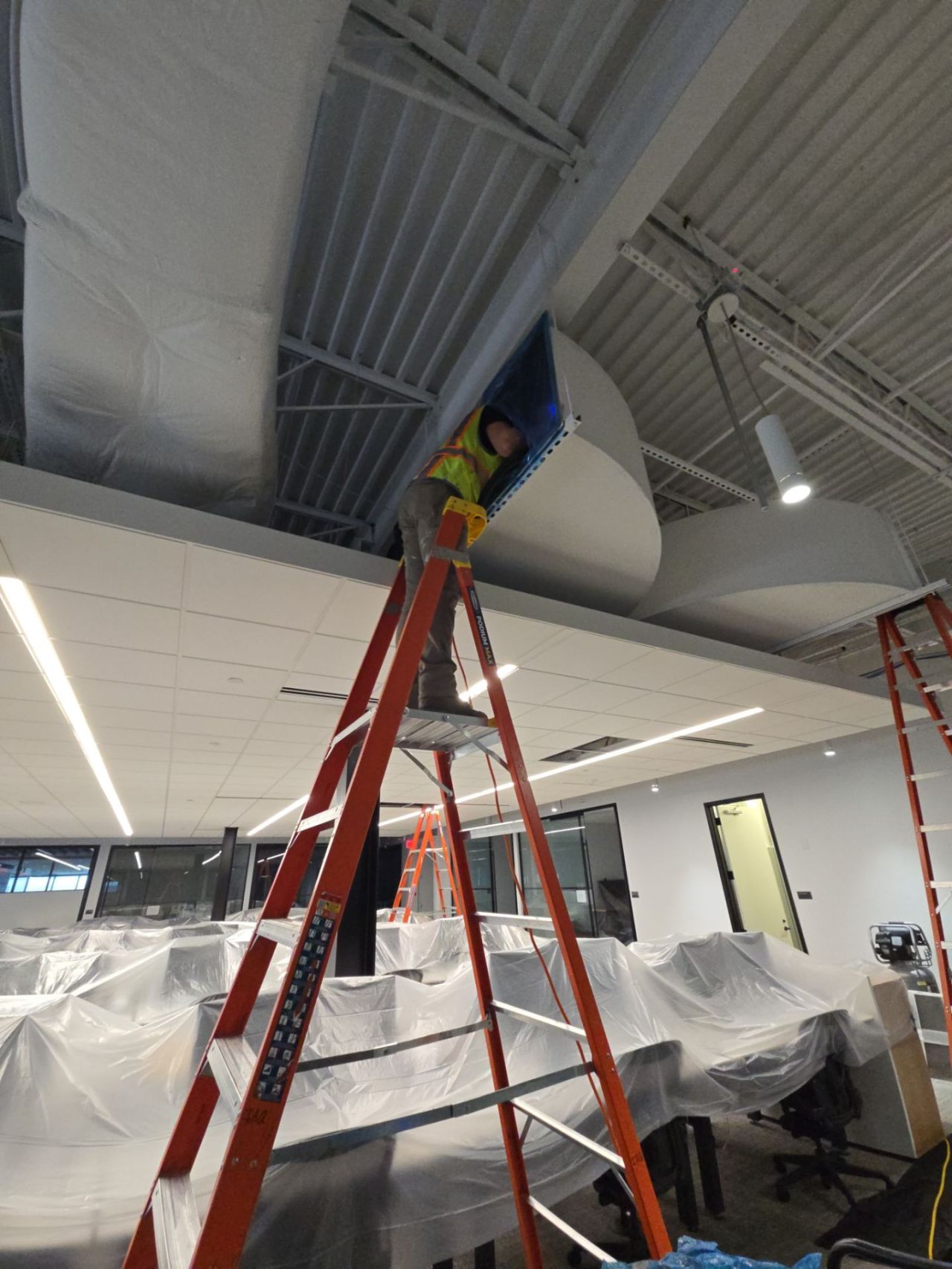 IAQ Restoration technician cleaning overhead ductwork above a fully wrapped, occupied office under containment